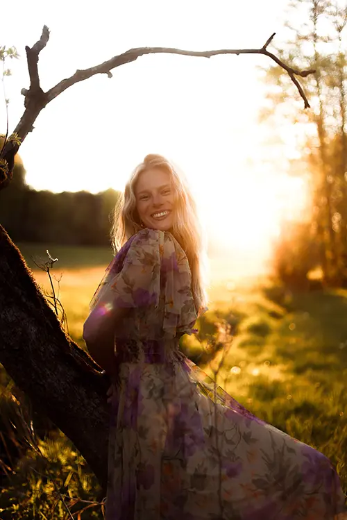 Blonde Frau in geblümtem Kleid lächelt im Sonnenlicht auf einem Feld.
