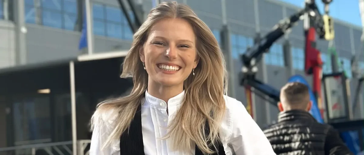 Lächelnde Frau mit langen blonden Haaren, weißer Bluse und Ohrring vor unscharfem Hintergrund, Nasenspitze hat eine kleine weiße Creme.
