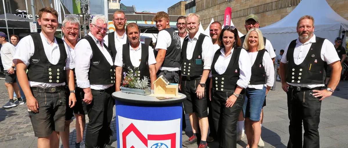 Drei Männer in traditioneller Handwerkerkleidung stehen draußen an einem runden Tisch mit einem kleinen Holzmodellhaus.