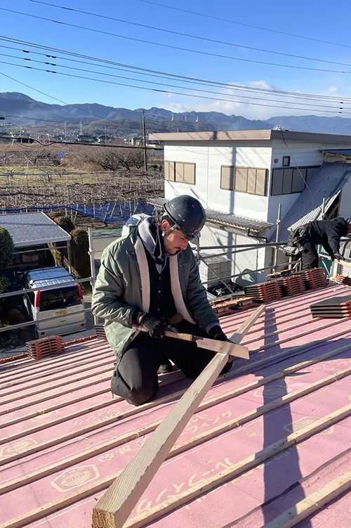 Ein Arbeiter mit Schutzhelm kniet auf einem Dach und arbeitet an Holzbalken unter blauem Himmel und vor einem weißen Gebäude.