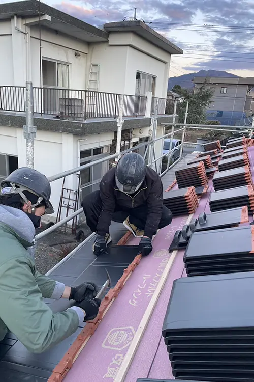 Eine Person mit Helm arbeitet auf einem Dach, verwendet Werkzeuge und baut oder repariert Dachziegel. Im Hintergrund ist ein Gebäude mit Balkon zu sehen.