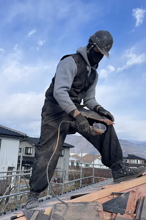 Eine Person mit Schutzbrille und Handschuhen arbeitet draußen mit einem Werkzeug auf einem Dach, im Hintergrund Häuser und blauer Himmel.