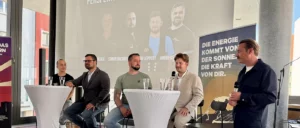 Ein Mann sitzt an einem Tisch mit zwei Gläsern Wasser, hinter ihm ist ein Bild mit zwei Männerporträts und Namen im Hintergrund projiziert.