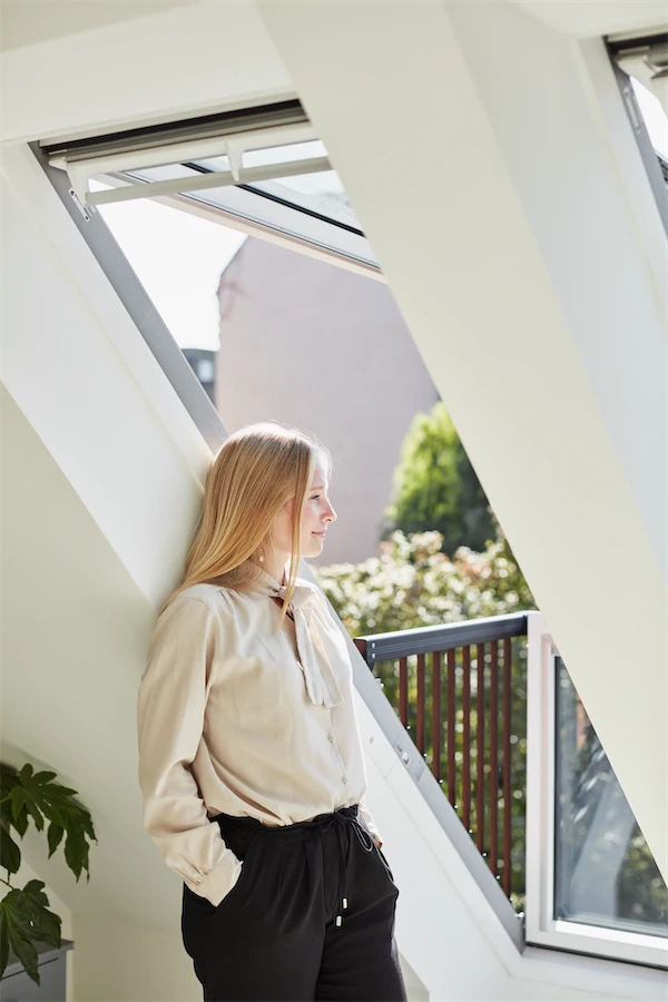 VELUX Markenprofil Eine Frau steht auf einem Balkon und blickt entspannt nach draußen, mit Sonnenlicht im Hintergrund.