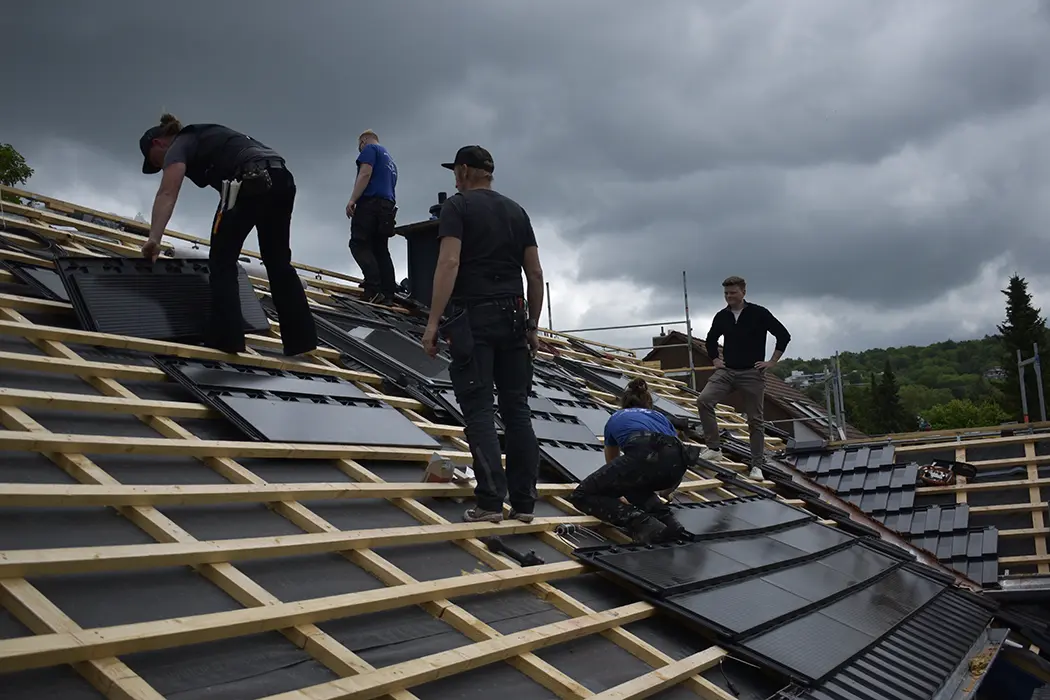 Ein Arbeiter montiert Solarmodule auf einem Hausdach unter bewölktem Himmel.