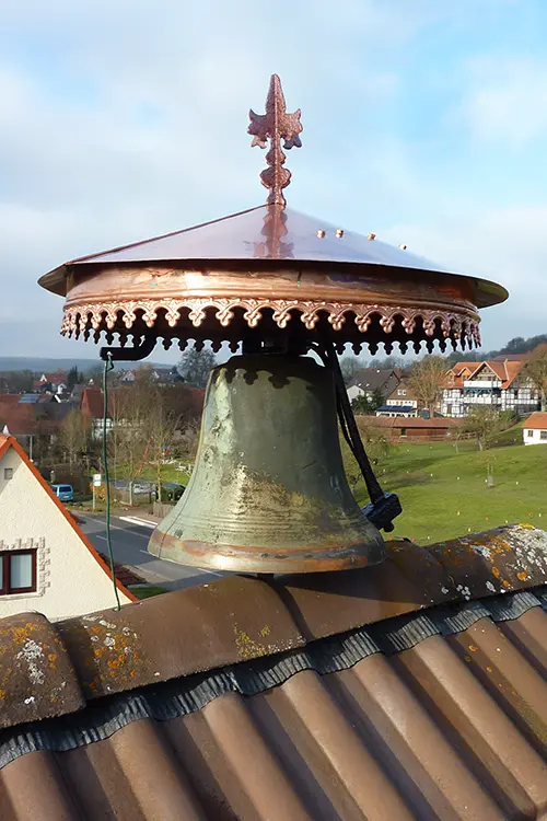Große Kirchenglocke aus Metall unter einem verzierten Kupferdach, im Freien auf einem Gebäudedach montiert, bei Tageslicht fotografiert.