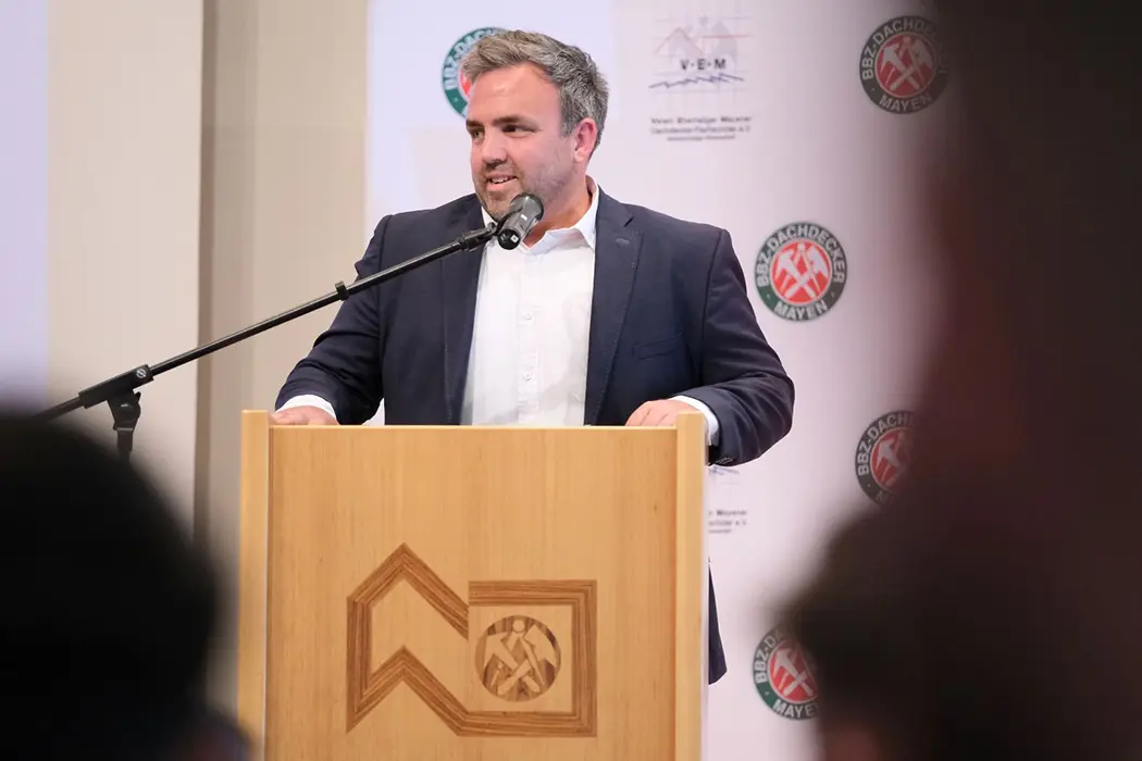 Ein Mann im Anzug spricht an einem Rednerpult mit Mikrofon, Symbol einer Organisation auf der Vorderseite des Pults.