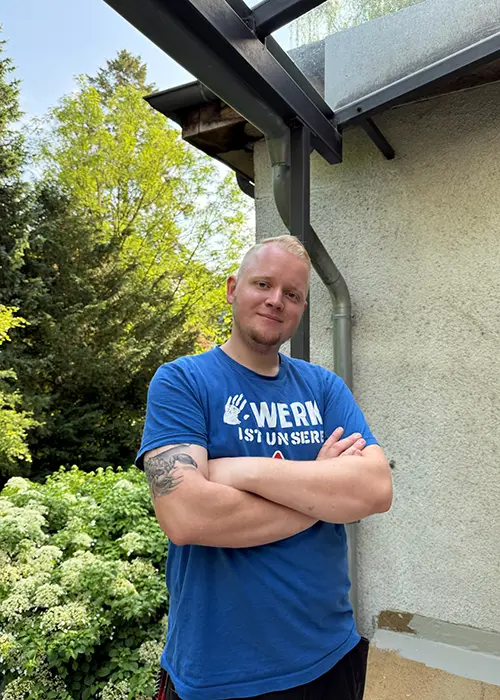 Ein Mann mit kurzen Haaren steht draußen, trägt ein blaues T-Shirt und hat die Arme verschränkt. Im Hintergrund sind Bäume und ein Teil eines Hauses zu sehen.