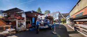 Ein Arbeiter steht neben einer großen blauen Maschine auf einem Lastwagen, Bäume und ein Hausdach sind im Hintergrund sichtbar.