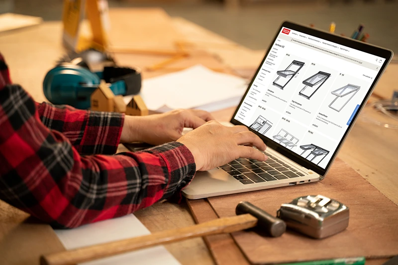VELUX Preiskatalog Hände tippen auf einem Laptop auf einem Holztisch, im Hintergrund liegen Papiere.