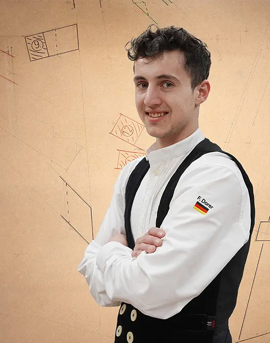 Junger Mann in traditioneller Kleidung mit verschränkten Armen, lächelt vor einem hellen Hintergrund mit Zeichnungen.