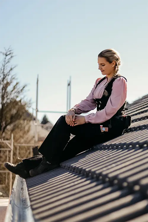 Eine Person sitzt auf einem Dach, trägt ein rosa Hemd und eine Armbanduhr, Hände ruhen auf den Knien, der Himmel ist klar.
