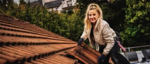 Eine Person mit blauen Handschuhen repariert Dachziegel auf einem Haus, im Hintergrund Bäume und ein Gebäude.