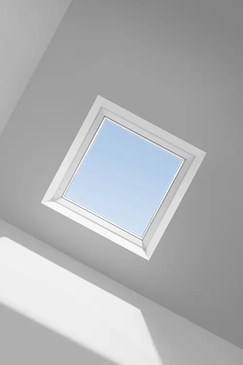 Ein rechteckiges Dachfenster mit weißem Rahmen zeigt einen klaren blauen Himmel, Licht fällt in einen weißen Raum.