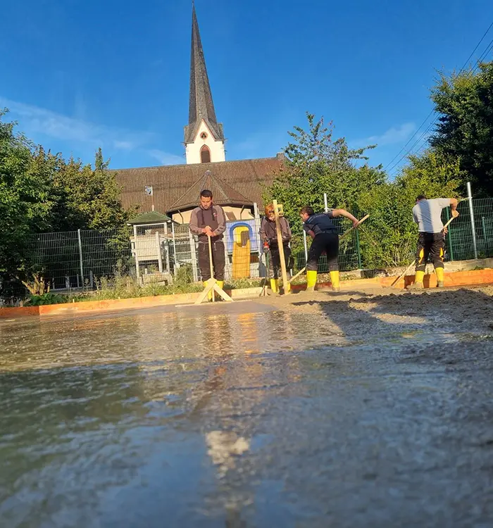 Zwei Bauarbeiter ebnen eine frisch gegossene Betonfläche vor einer Kirche bei sonnigem Wetter.