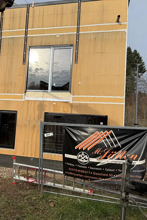Fenster in einem unfertigen Holzbau, Bauzaun mit Werbebanner einer Zimmerei im Vordergrund, draußen bei Tageslicht.