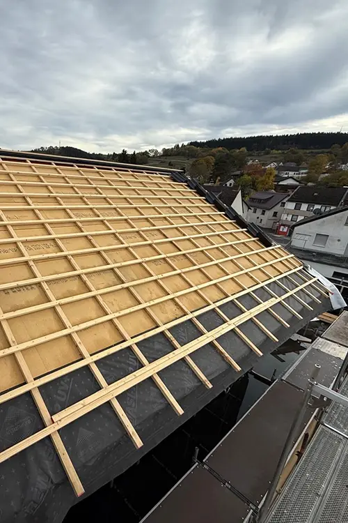 Ein Dach im Bau mit Holzlatten auf einer schwarzen Abdichtung, darunter bewölkter Himmel und bewaldete Hügel im Hintergrund.