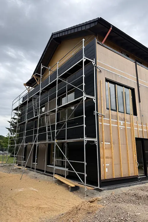 Ein Baugerüst steht an einer schwarzen Hausfassade, offenbar für Bau- oder Renovierungsarbeiten, bei bedecktem Himmel.