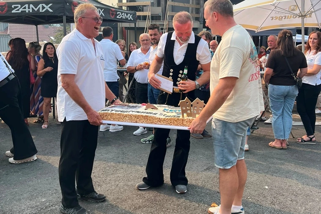 Zwei Männer stehen im Freien und tragen eine Torte, Arno Billen trägt traditionelle Kleidung und schaut auf den Kuchen, im Hintergrund sind viele Gäste der 25-Jahr-Feier des Betriebs zu sehen.