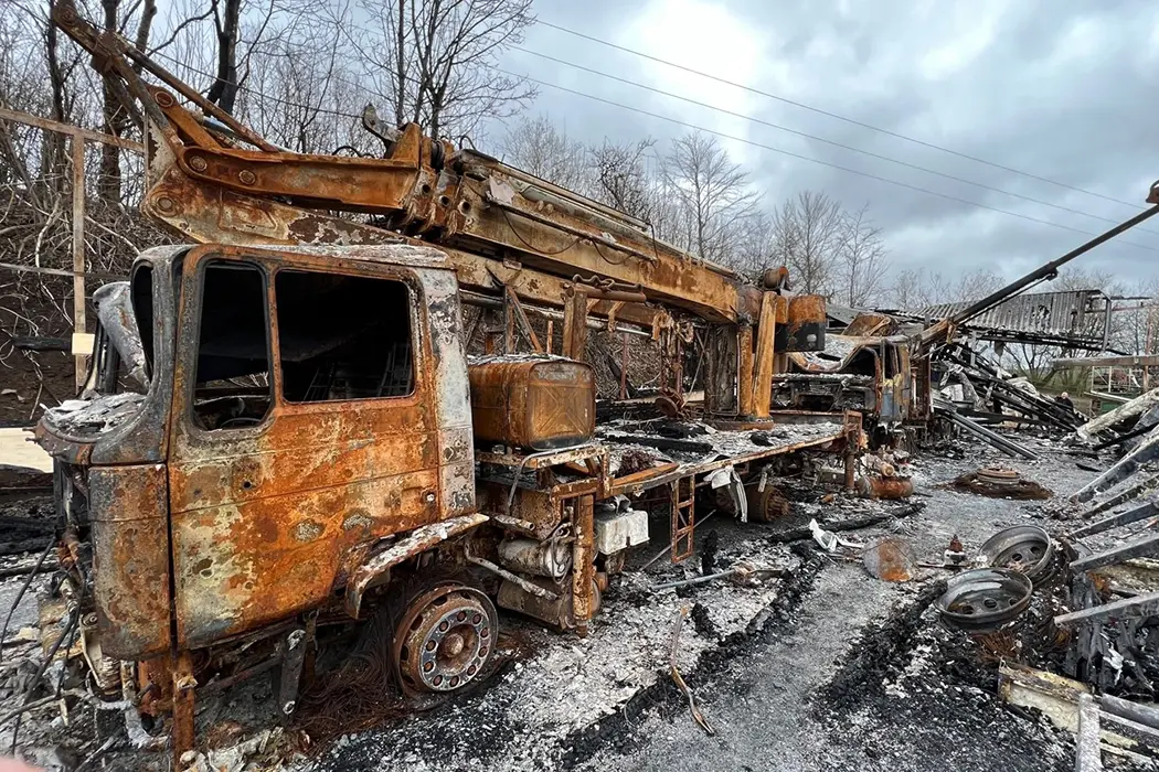 Verrostete Industriemaschine im Freien, umgeben von trockenem Boden und kahlen Bäumen an einem bewölkten Tag.