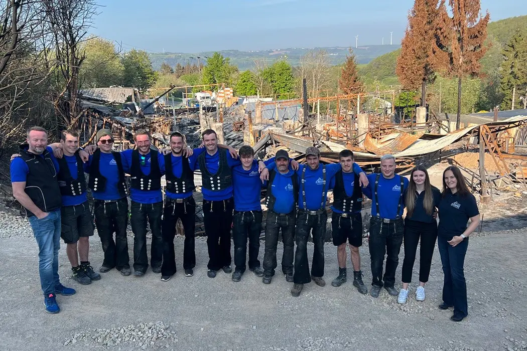 Zwei Bauarbeiter in blauen Pullovern stehen Arm in Arm vor einer Baustelle mit Trümmern und Bäumen im Hintergrund.