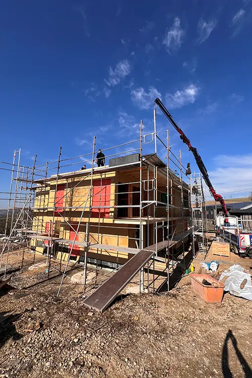 Eine Baustelle mit eingerüstetem Haus, blauen Himmel und einer Person, die auf dem Dach arbeitet. Baustellenmaterialien und eine Rampe sind im Vordergrund sichtbar.