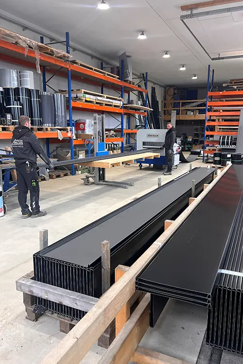 Werkstatt mit Regalen voller Materialien, einer Werkbank, Maschinen und Metallprofilen im Vordergrund. Industrielle Beleuchtung von der Decke, aufgeräumte und strukturierte Umgebung.