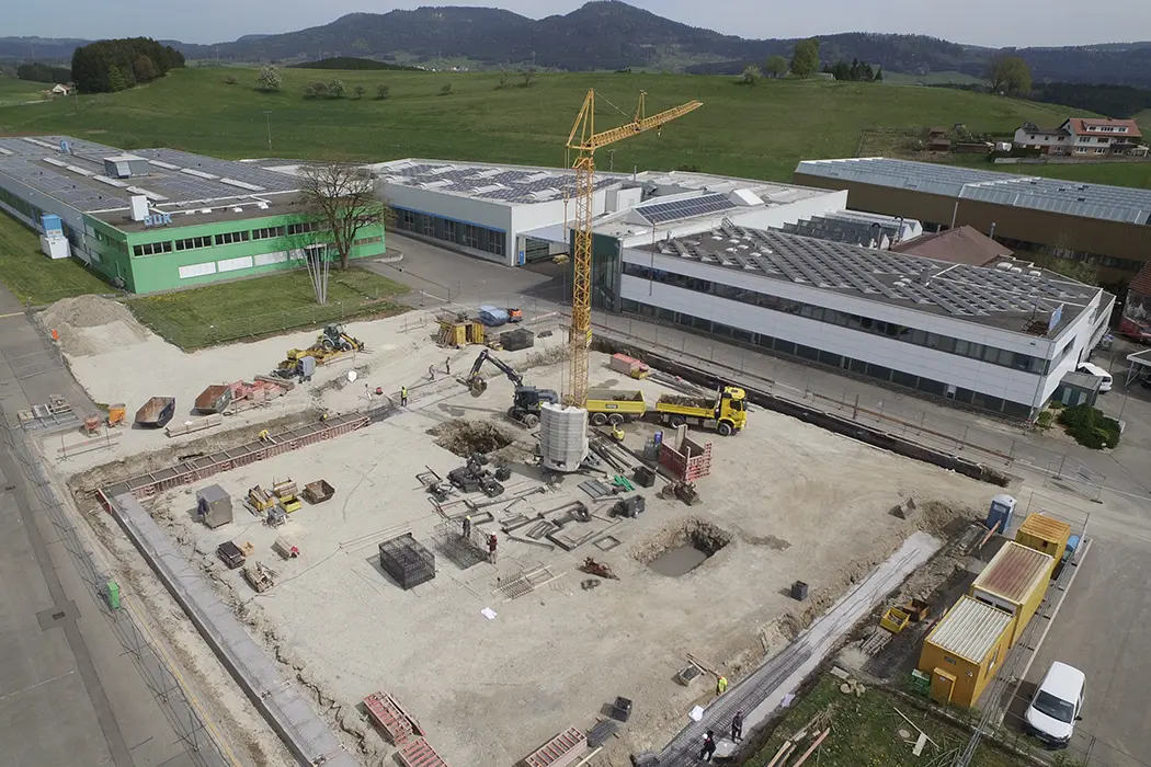Baustelle mit gelbem Kran, Baumaterialien und Maschinen, im Hintergrund grüne Wiesen, Berge und Industriegebäude.