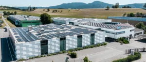 Moderne Gebäude mit Solarpaneelen auf dem Dach, einer weißen Fassade, grüne Pflanzen im Vordergrund und hügelige Landschaft im Hintergrund bei Tageslicht.