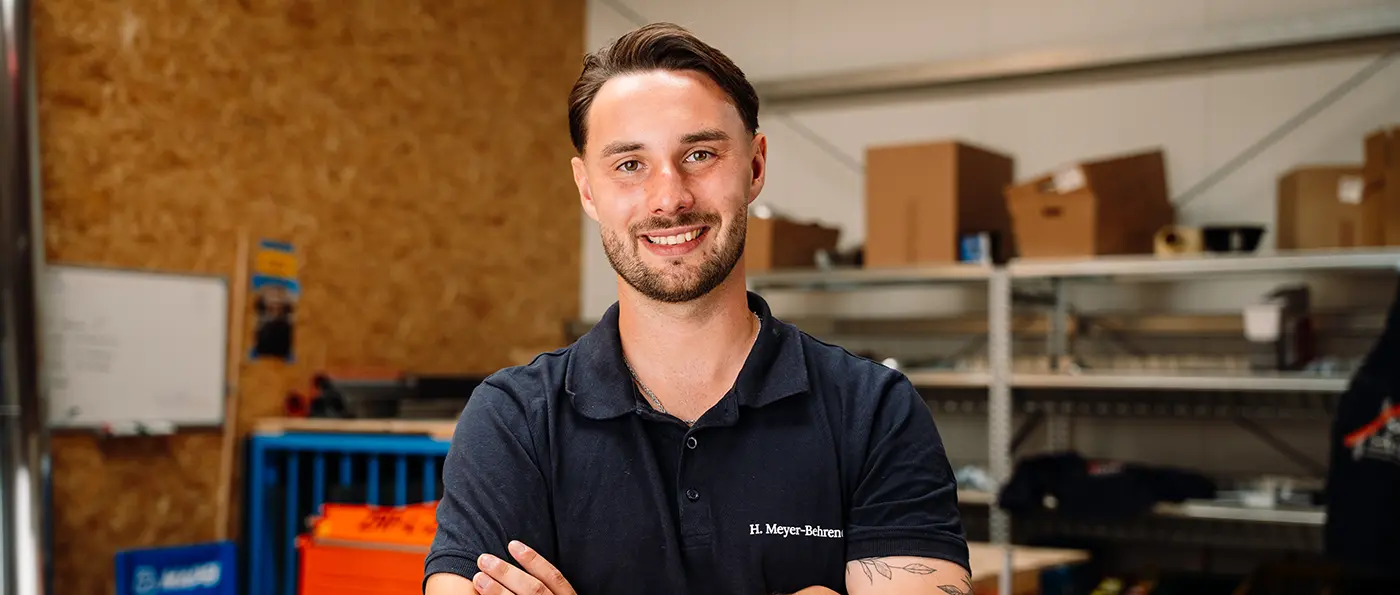 Ein junger Mann mit Bart und kurzem Haar lächelt, trägt ein dunkelblaues Poloshirt, Hintergrund unscharf in Innenraum.