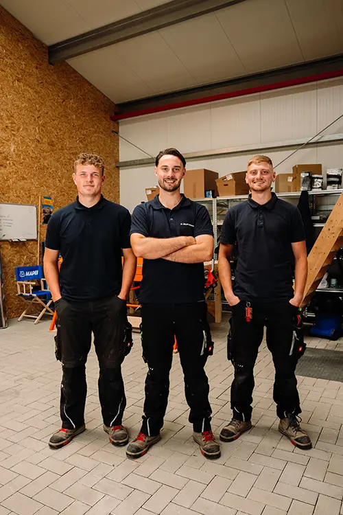Ein Mann steht in einer Werkstatt mit verschränkten Armen und lächelt, trägt Arbeitskleidung und Turnschuhe, umgeben von Regalen und Kartons im Hintergrund.