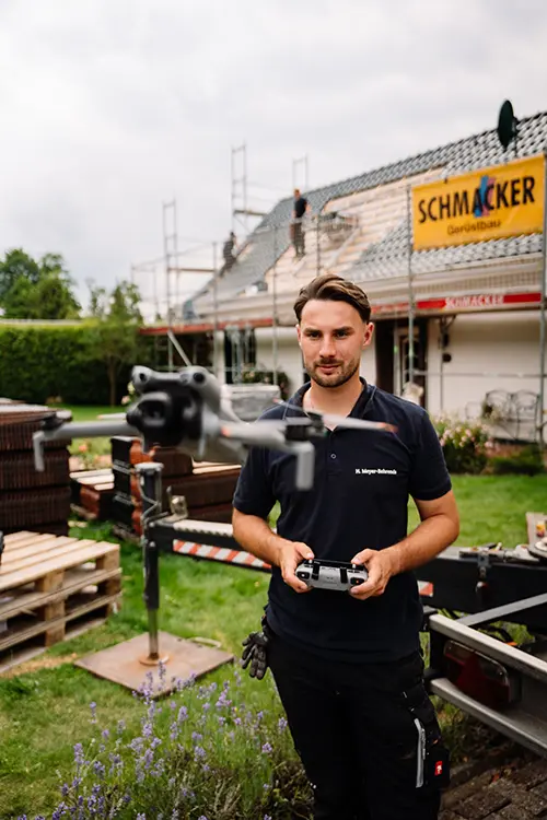 Ein Mann steuert eine Drohne vor einem Haus, auf dessen Dach Arbeiter arbeiten.