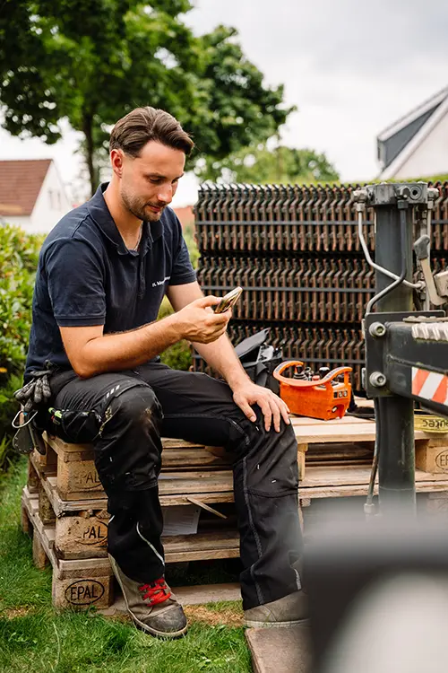 Ein Mann sitzt draußen, trägt Arbeitskleidung und schaut auf sein Smartphone, Baumaterialien und Werkzeuge im Hintergrund.