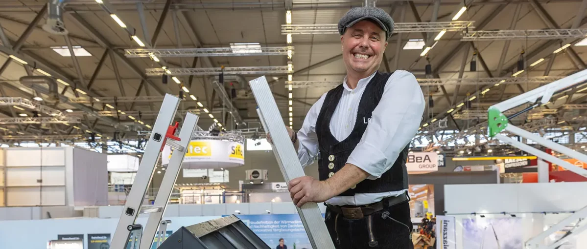 Eine Person hält ein langes rechteckiges Holzbrett in einer Messehalle, Hintergrund zeigt Decke mit Lichtern und einen verschwommenen Informationsstand.