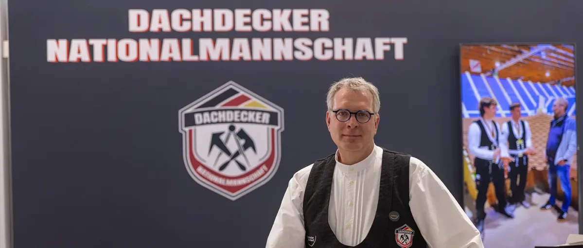 Teilweise sichtbares Logo und weiße Buchstaben auf dunklem Hintergrund, ein Teil einer Person mit weißem Hemd und schwarzer Weste am rechten Bildrand.