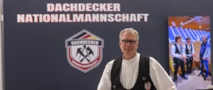 Teilweise sichtbares Logo und weiße Buchstaben auf dunklem Hintergrund, ein Teil einer Person mit weißem Hemd und schwarzer Weste am rechten Bildrand.