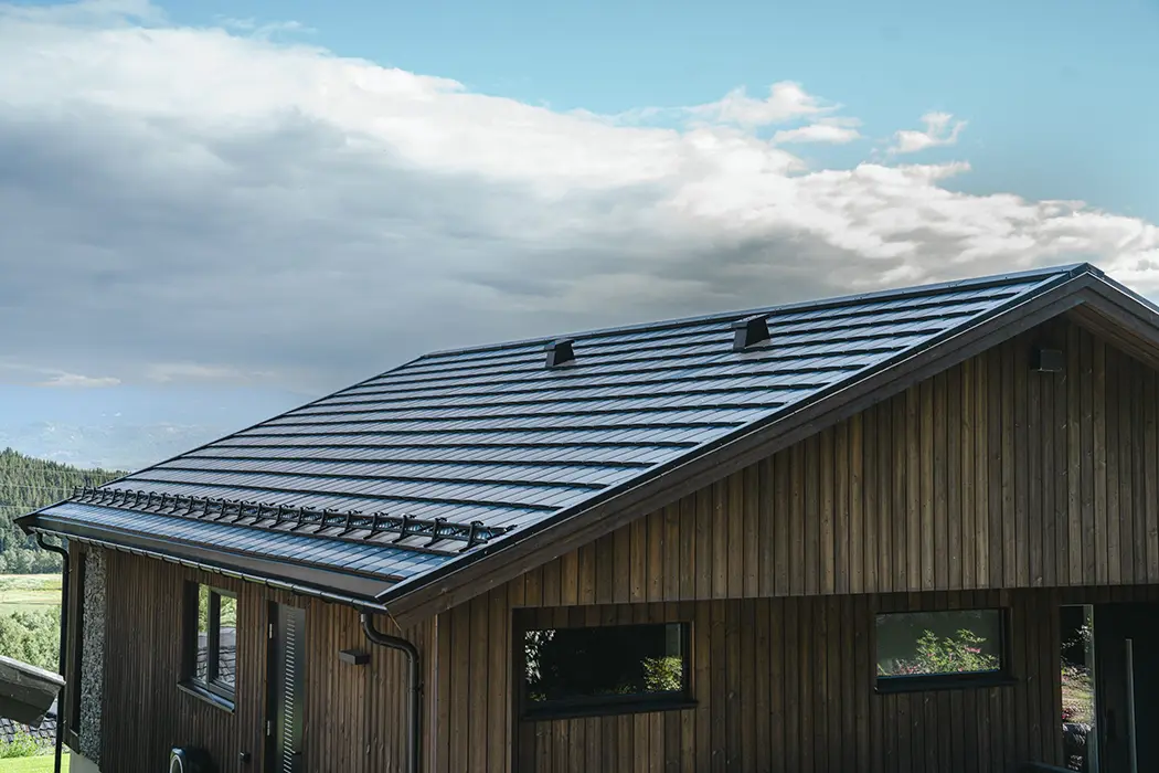 Schrägdach mit dunkel glänzenden Solardachziegeln auf einem modernen Holzhaus, blauer Himmel und einige Wolken im Hintergrund.
