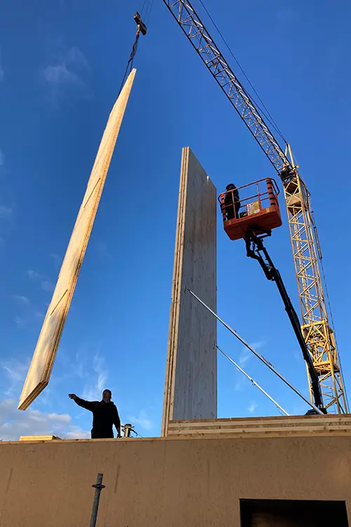 Eine große Betonwand wird mit einem Kran auf einer Baustelle aufgerichtet, klarer blauer Himmel im Hintergrund.