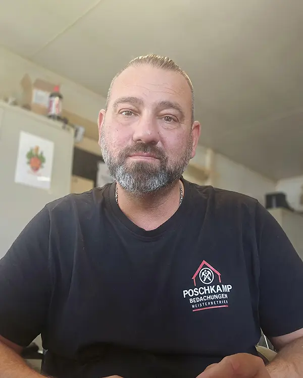 Ein Mann mit Bart und grauen Haaren trägt ein schwarzes T-Shirt und schaut direkt in die Kamera.
