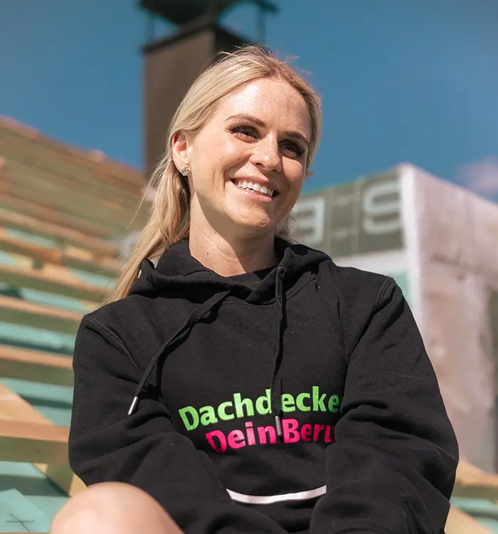 Eine lächelnde Frau mit blondem Haar trägt einen schwarzen Hoodie mit bunter Aufschrift, im Freien bei Sonnenschein.