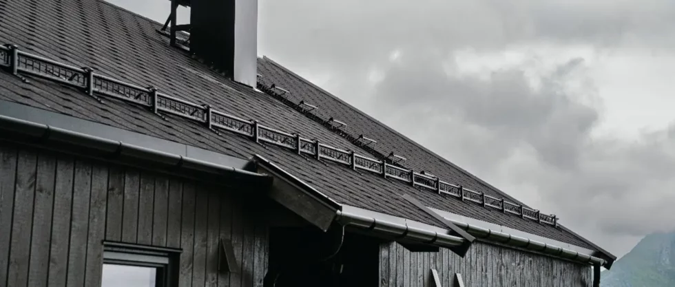 Schräge Dachfläche eines Gebäudes mit dunklen Dachziegeln und Schneefanggittern, unter bewölktem Himmel.