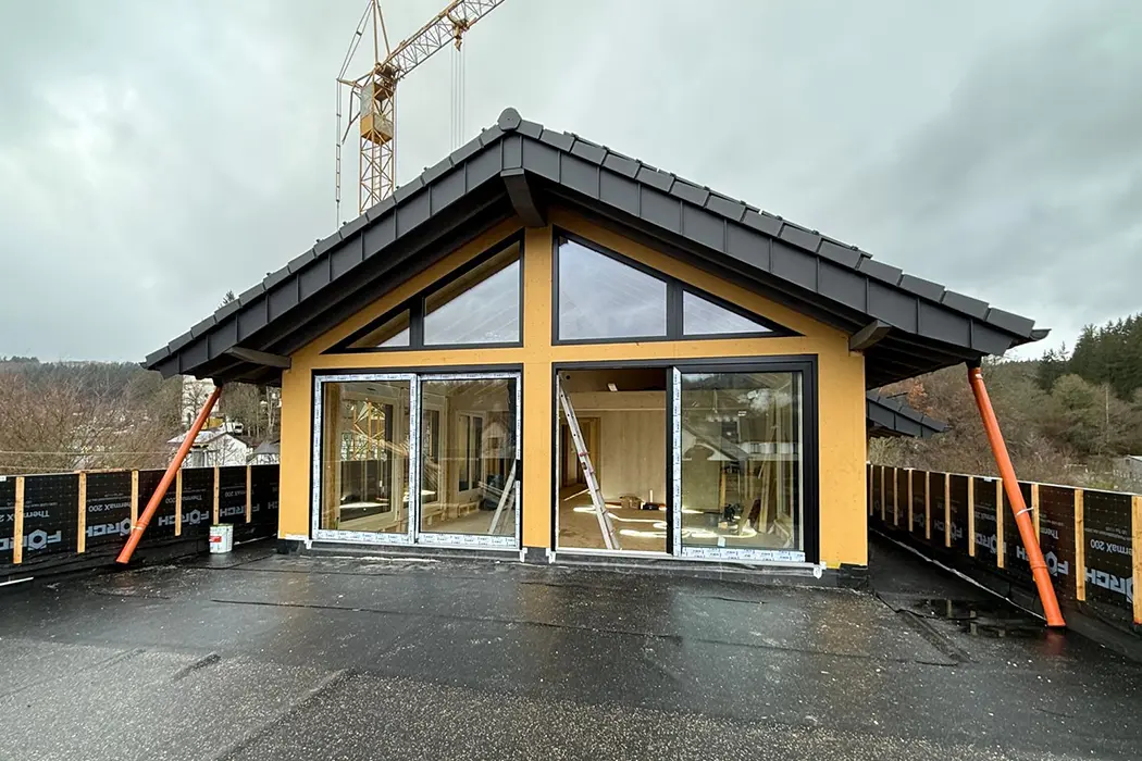 Teil eines modernen Holzhauses mit großen Fenstern, gelbem Rahmen und schwarzem Dach, innen eine Leiter sichtbar, draußen nasser Asphalt.