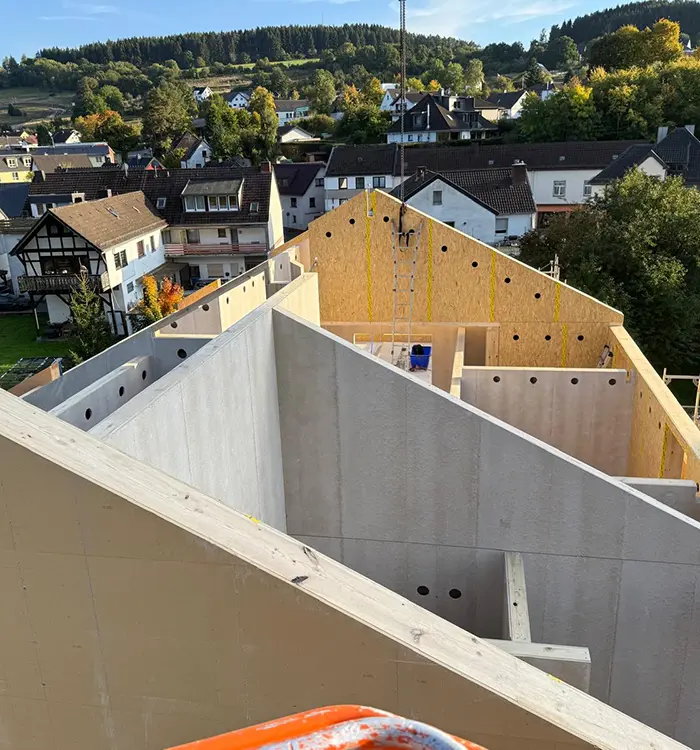 Baustelle mit Beton- und Holzstrukturen, im Hintergrund Wohnhäuser und Bäume unter blauem Himmel.
