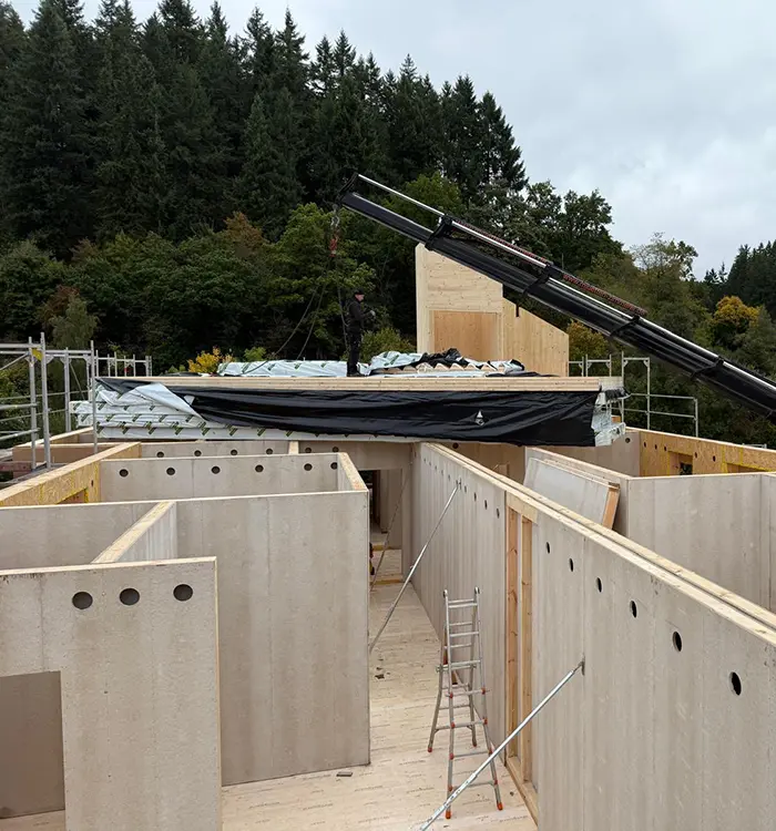 Baustelle mit Holzrahmen, Kran, Bauarbeiter auf dem Dach und Wänden im Bau, umgeben von Bäumen im Hintergrund.