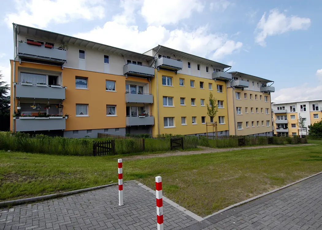 Mehrstöckiges gelbes Wohnhaus mit Balkonen, davor eine Wiese, ein kleiner Zaun und ein rot-weiß gestreifter Pfosten auf dem Gehweg.
