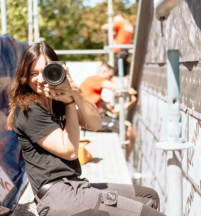Zwei Personen arbeiten im Freien an einer Baustelle, eine davon im Vordergrund teilweise zu sehen, die andere im Hintergrund, beide tragen orangefarbene Kleidung.