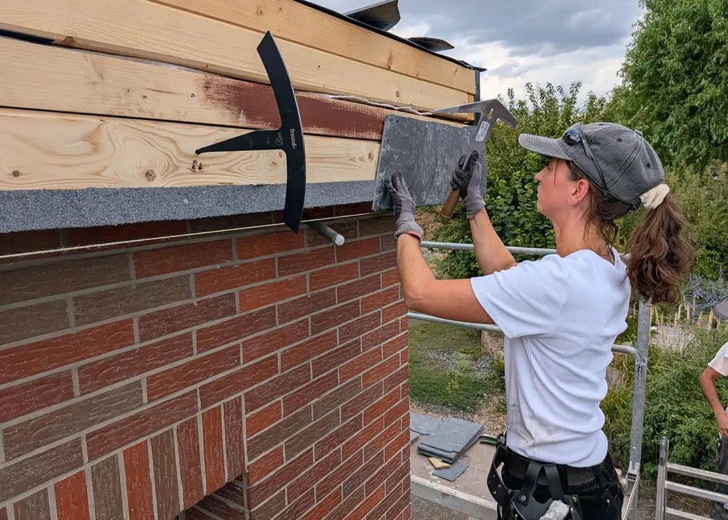 Eine Person mit Handschuhen arbeitet an einer Dachkante aus Holz und Ziegeln, hält eine Schieferplatte an die Wand.
