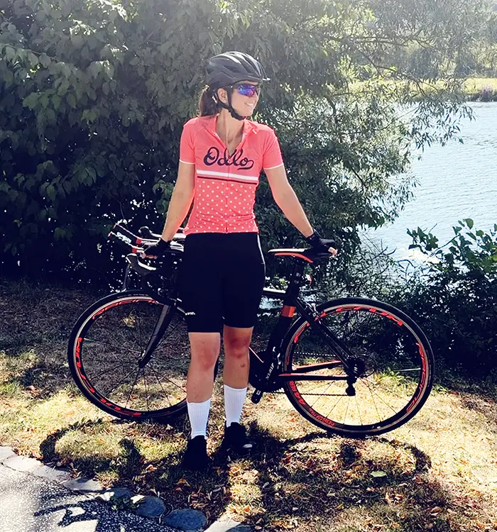 Frau mit Fahrradhelm, Sonnenbrille und pinkem Radtrikot steht neben einem Fahrrad im Freien.