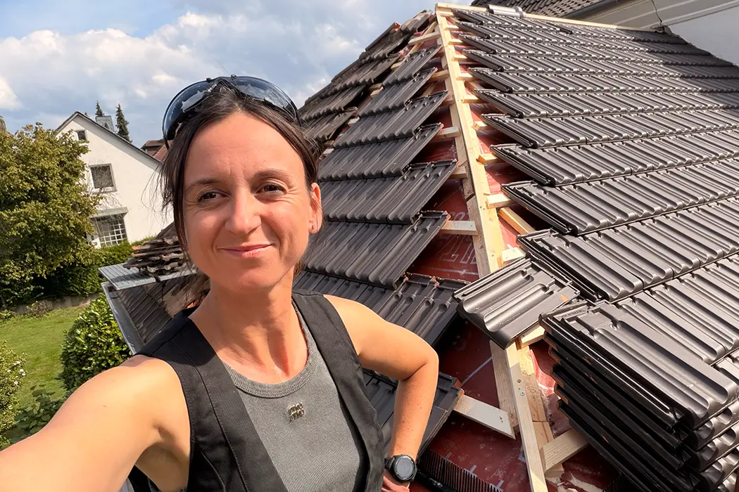Ein Teil einer Person mit schwarzer Weste steht vor einem geneigten Hausdach mit dunklen Dachziegeln.