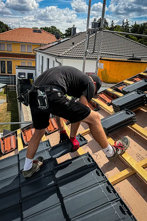 Ein Arbeiter in schwarzer Kleidung und roten Handschuhen verlegt Dachziegel auf einem Hausdach an einem sonnigen Tag.